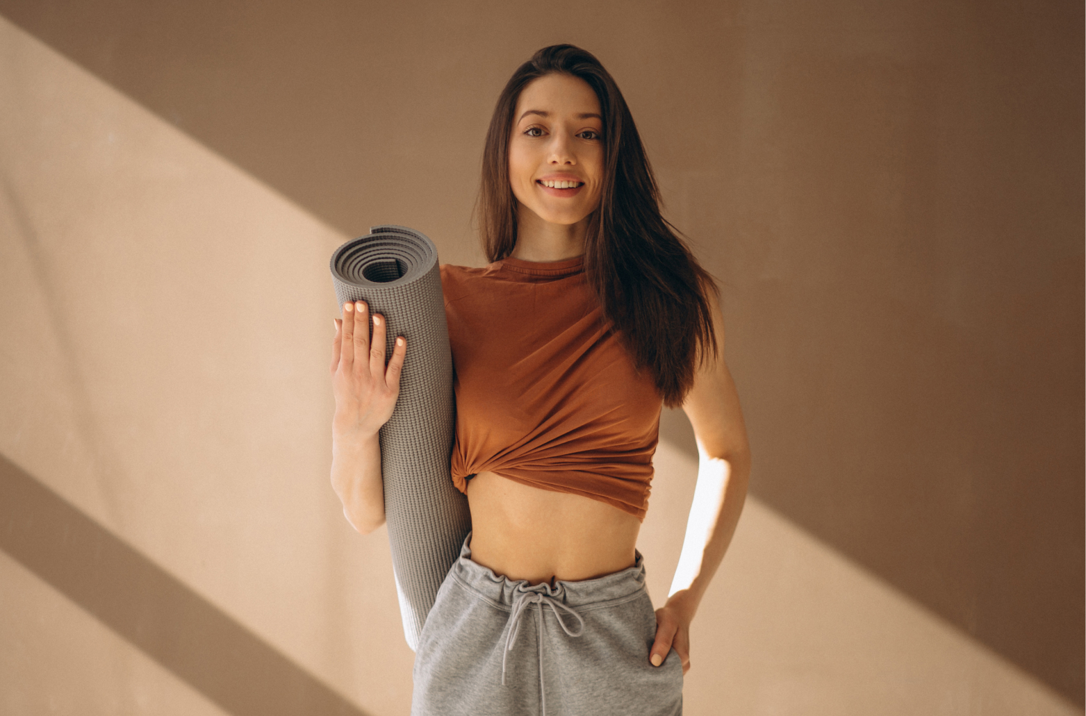 Молодая женщина в спортивной одежде с ковриком для йоги в руках, улыбается и стоит в светлом интерьере, демонстрируя здоровый и активный образ жизни
